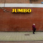 een oudere vrouw loopt voor een bakstenen muur met daarop het logo van supermarktketen Jumbo