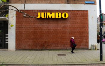 een oudere vrouw loopt voor een bakstenen muur met daarop het logo van supermarktketen Jumbo