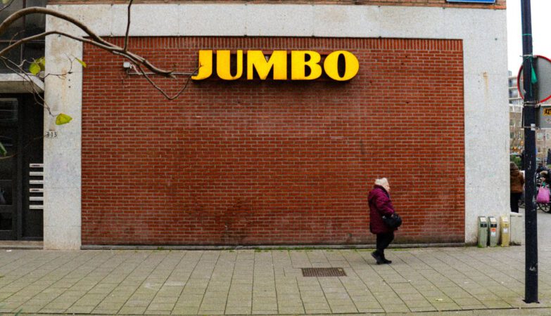 een oudere vrouw loopt voor een bakstenen muur met daarop het logo van supermarktketen Jumbo